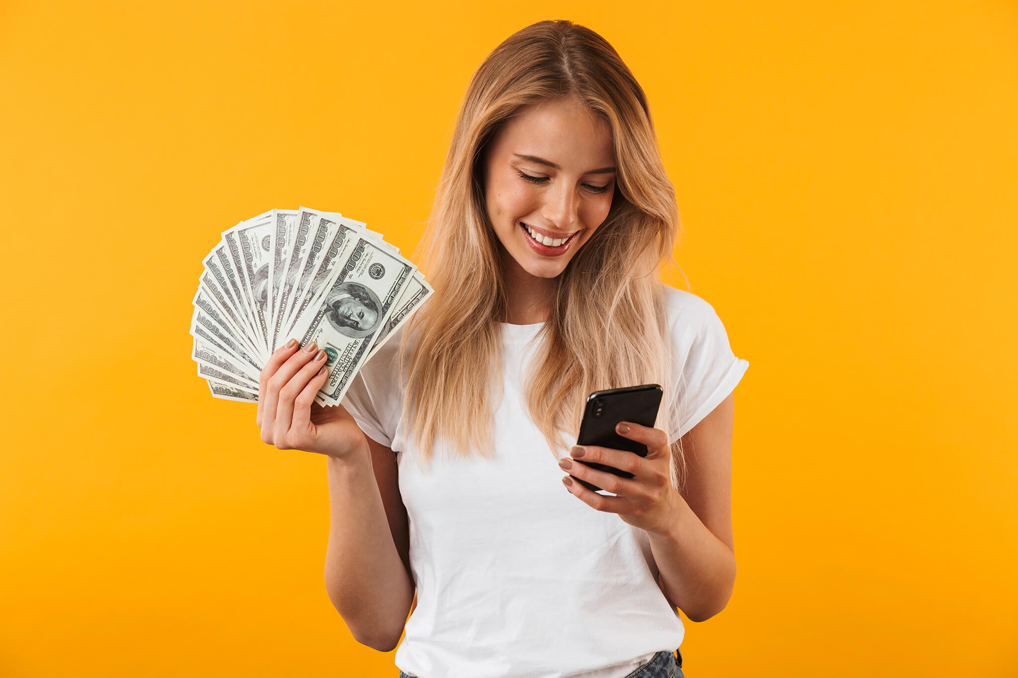 A woman with money in her right hand looking at her smartphone in her left hand